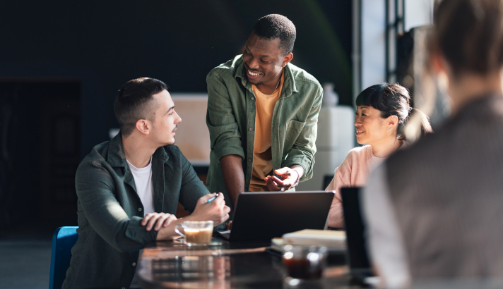 Man standing over two people talking around a laptop.