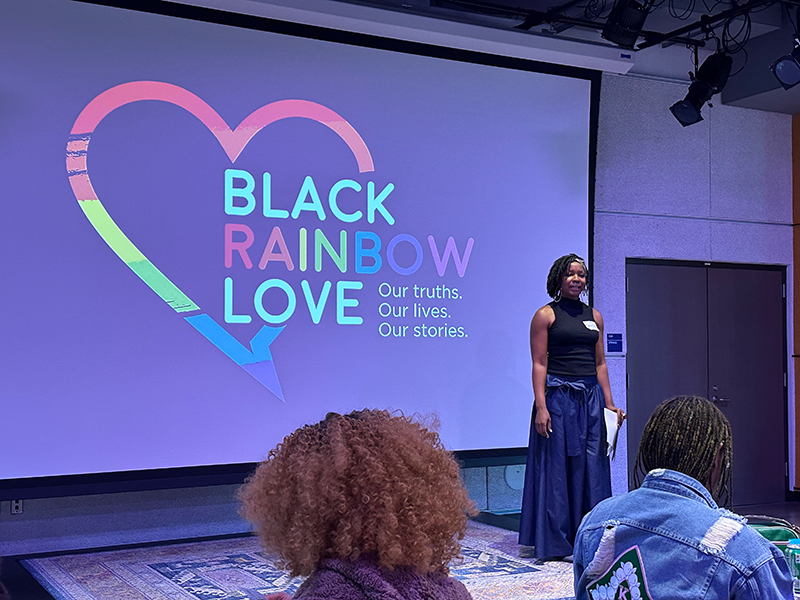Woman presenting in front of a screen that says "Black Rainbow Love. Our truths. Our lives. Our stories."
