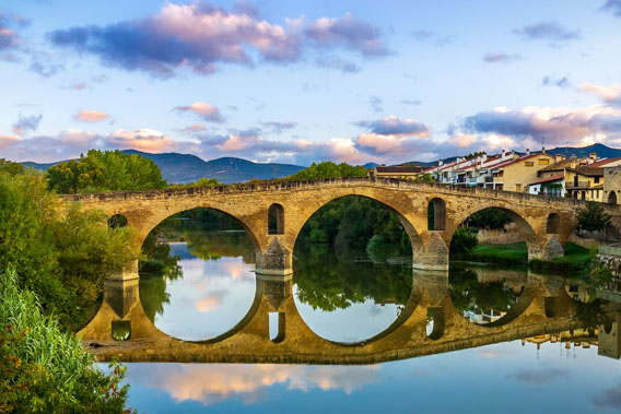 Old stone bridge over calm river with town on the right and lush trees on the left