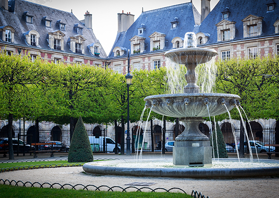 Large water fountain in town square surrounded by trees and buildings in background